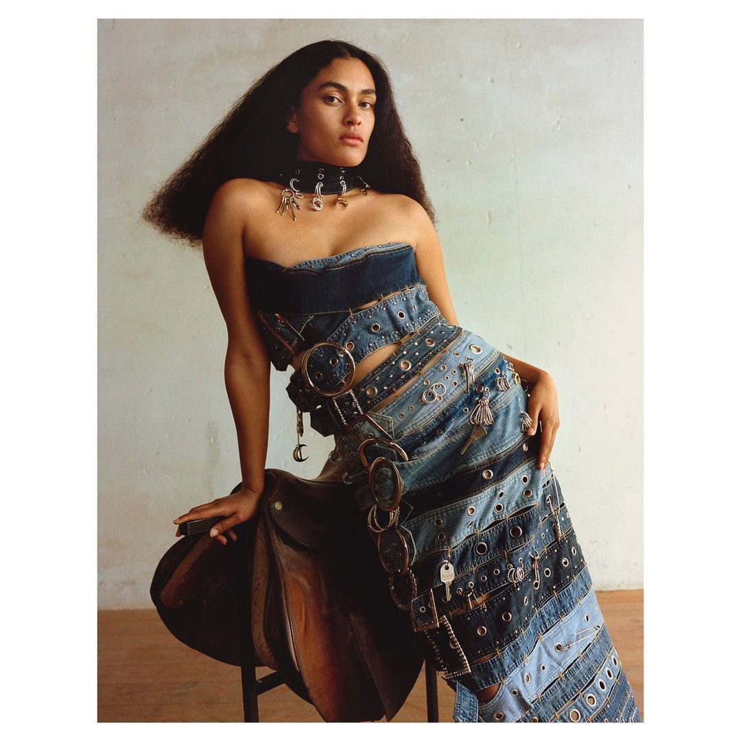 A striking fashion shot of an adult woman with long dark hair in a strapless denim dress, posing with one hand on a brown saddle.