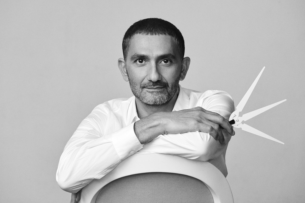 Portrait of a middle-aged man in a white shirt sitting on a white chair, holding a drafting tool in his hand, in a monochrome studio.