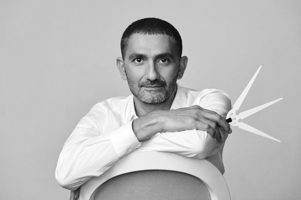 Portrait of a middle-aged man in a white shirt sitting on a white chair, holding a drafting tool in his hand, in a monochrome studio.