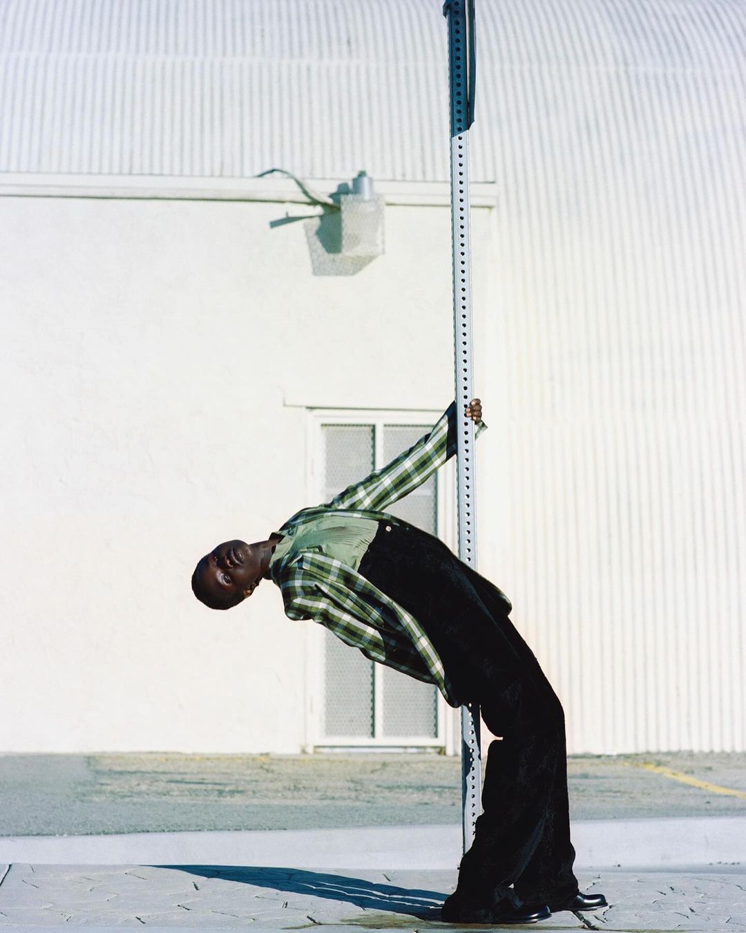 A black adult male bends backwards against a striped pole, wearing a plaid shirt and black pants, in an outdoor urban environment.