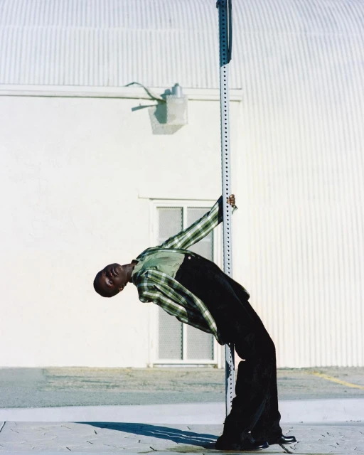 A black adult male bends backwards against a striped pole, wearing a plaid shirt and black pants, in an outdoor urban environment.