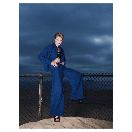 Model in blue matching jacket and wide-leg trousers standing on sand in front of a chain-link fence under a dark twilight sky.