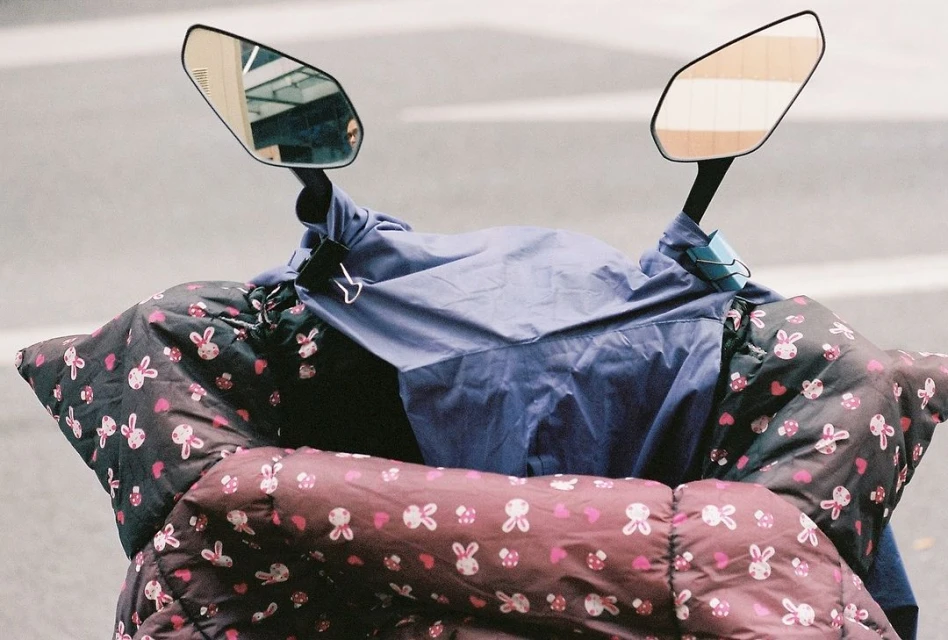 Motorcycle seat and mirrors with a blue cover and black, pink, and white patterned blanket over the seat, parked on a gray road.