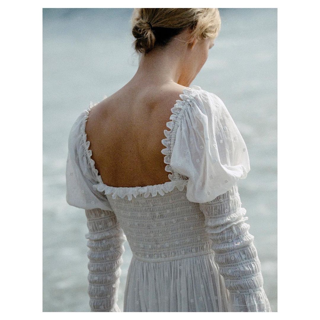 Woman in a white embroidered dress stands near the ocean, back view, with ruffled square neckline and long sleeves.