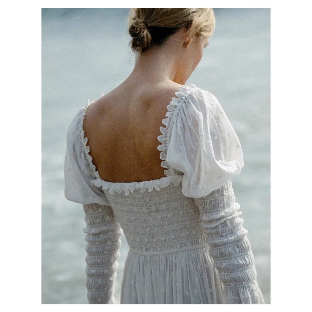 Woman in a white embroidered dress stands near the ocean, back view, with ruffled square neckline and long sleeves.