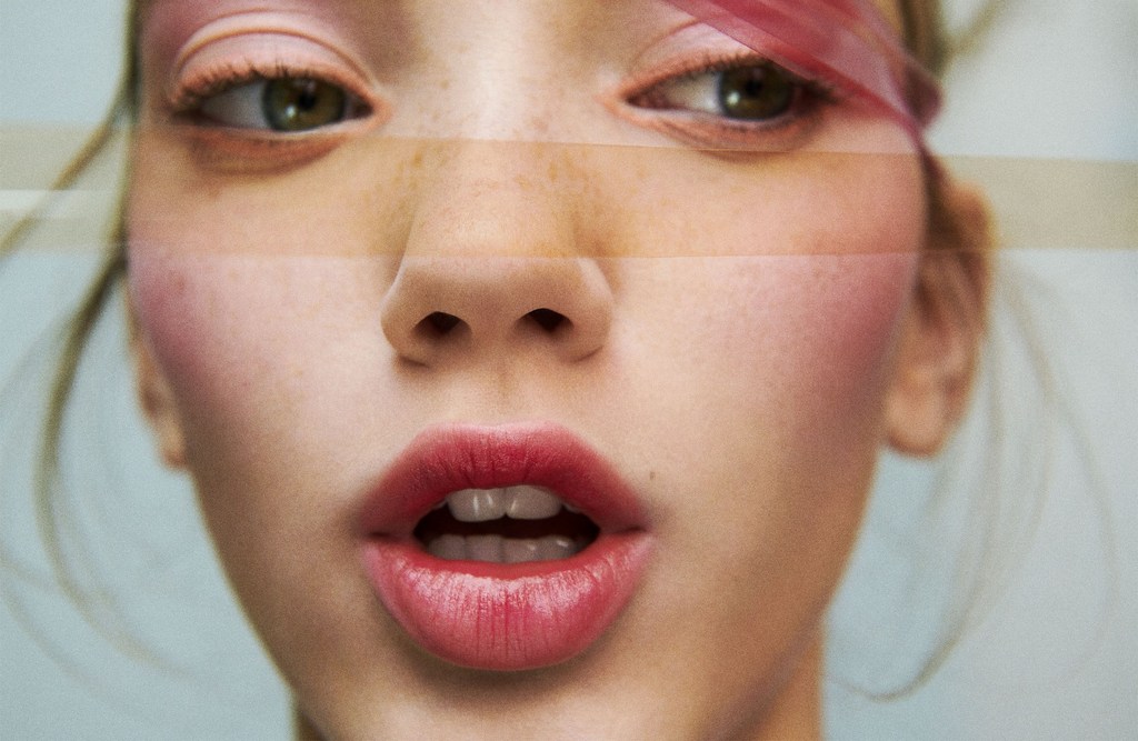 Close-up of a young adult female with fair skin, striking red lipstick, and light eye makeup, set against a soft blue background.