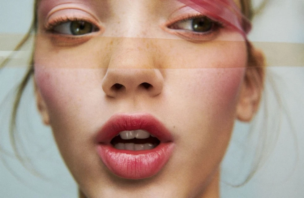 Close-up of a young adult female with fair skin, striking red lipstick, and light eye makeup, set against a soft blue background.