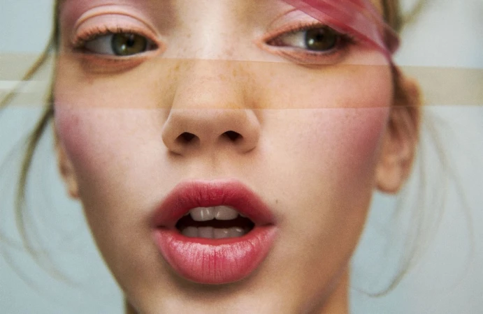 Close-up of a young adult female with fair skin, striking red lipstick, and light eye makeup, set against a soft blue background.