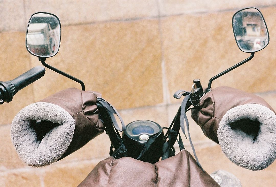 Close-up of a motorcycle handlebar with brown leather and gray fur-lined hand covers, black levers, and rearview mirrors against a tan wall.