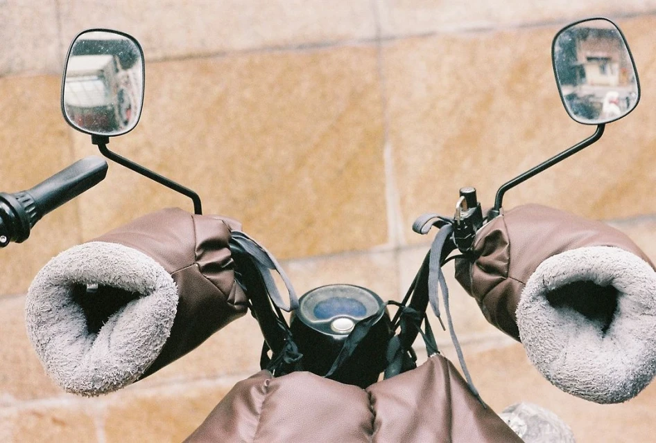Close-up of a motorcycle handlebar with brown leather and gray fur-lined hand covers, black levers, and rearview mirrors against a tan wall.