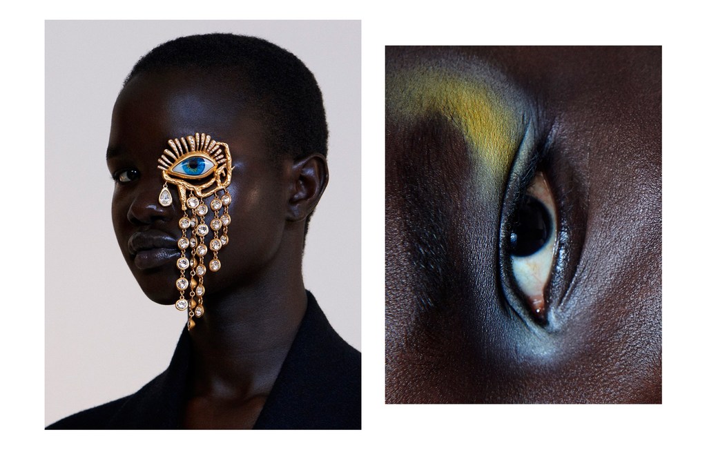Headshot of a woman wearing a large gold eye pendant earring with crystal fringe and makeup in a white studio setting.