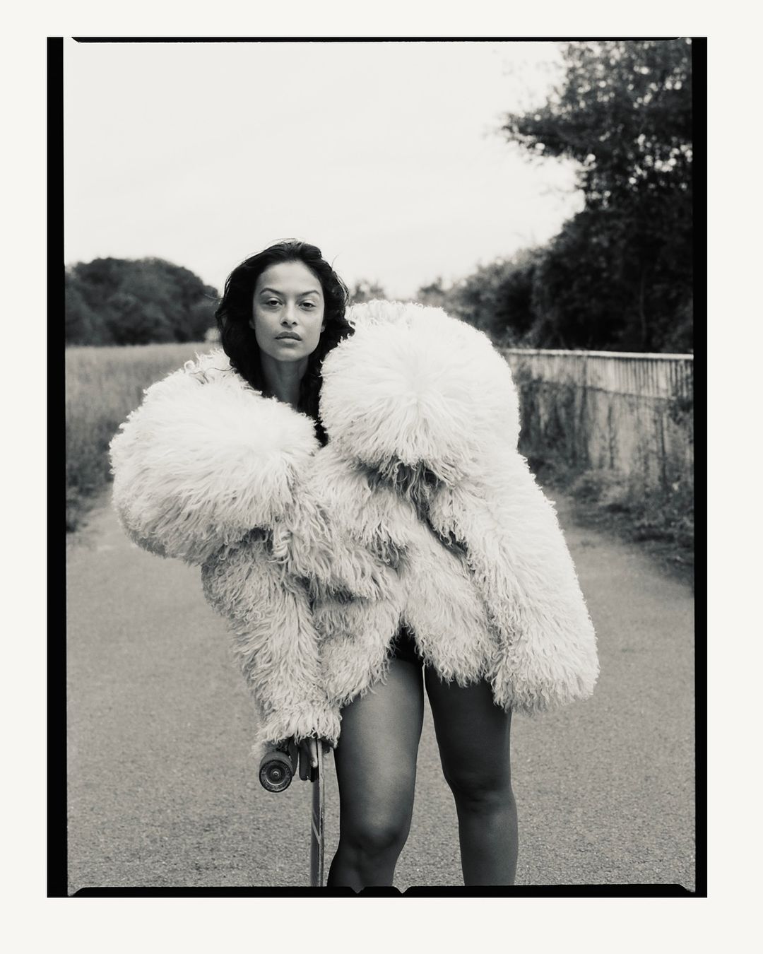 Fashionable woman with skateboard wearing a large fluffy coat on a road with vegetation. Moody black and white style portrait.
