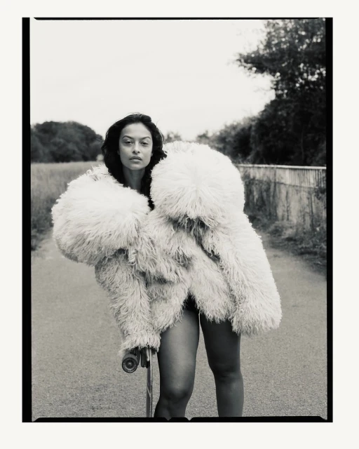 Fashionable woman with skateboard wearing a large fluffy coat on a road with vegetation. Moody black and white style portrait.