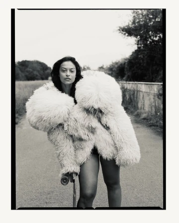 Fashionable woman with skateboard wearing a large fluffy coat on a road with vegetation. Moody black and white style portrait.