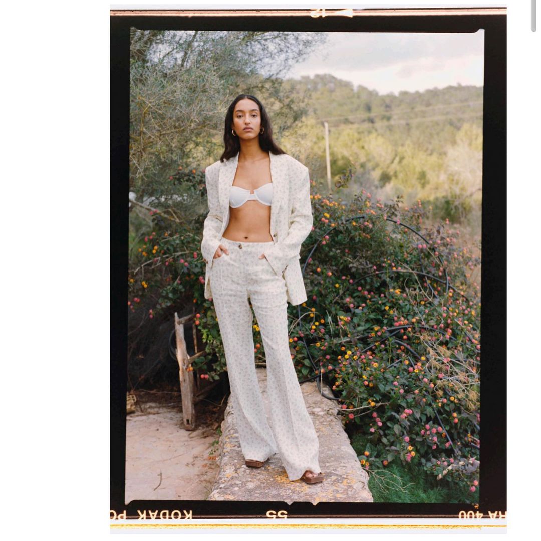 A brunette woman poses outdoors wearing a white blazer, white bra top, and white slacks, with a natural backdrop of trees and flowers.