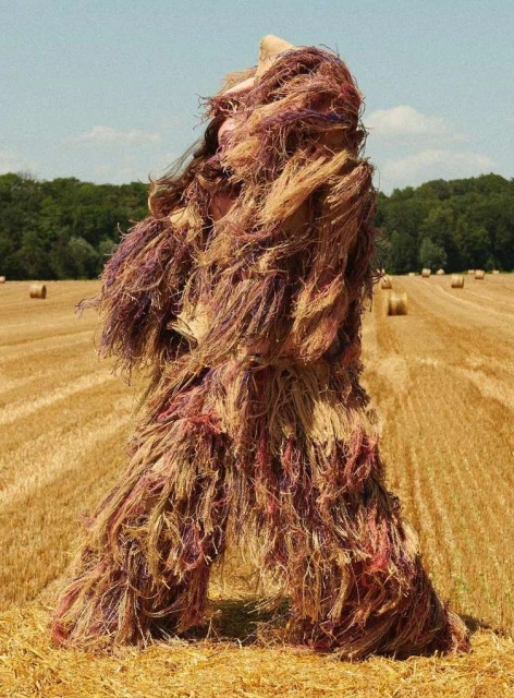 Person in fringed ghillie suit stands in an agricultural field with rolled hay bales under a blue sky on a sunny day.