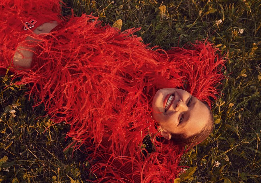 Woman in a red feathered coat lies on a grass field, smiling at the camera in a medium shot, capturing a blend of fashion and nature.