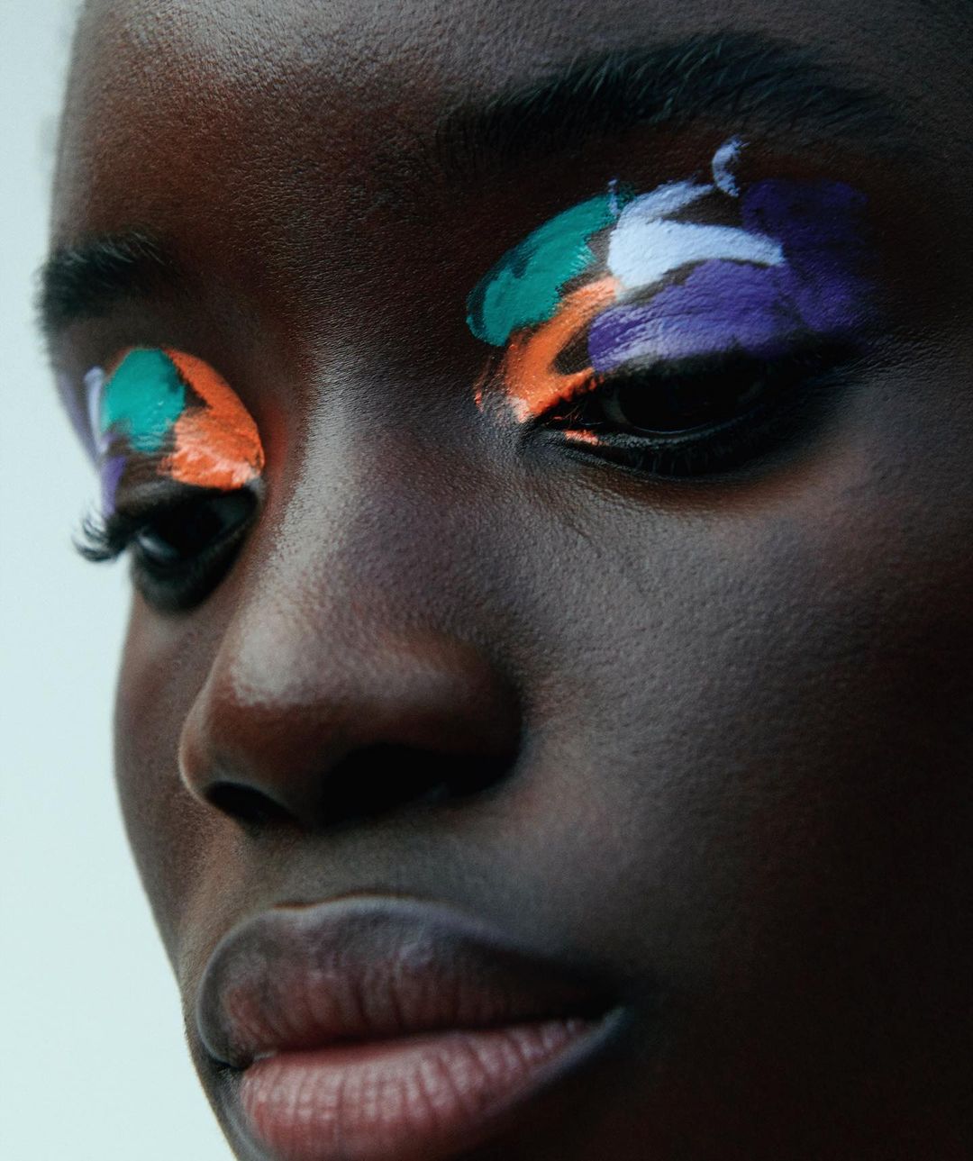 Close-up portrait of a dark-skinned female adult with colorful paint applied to her eyelids in hues of green, white, orange, and purple.