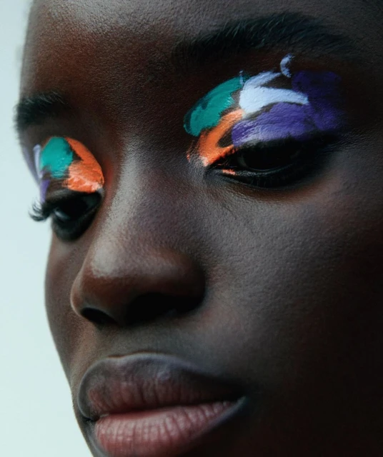 Close-up portrait of a dark-skinned female adult with colorful paint applied to her eyelids in hues of green, white, orange, and purple.