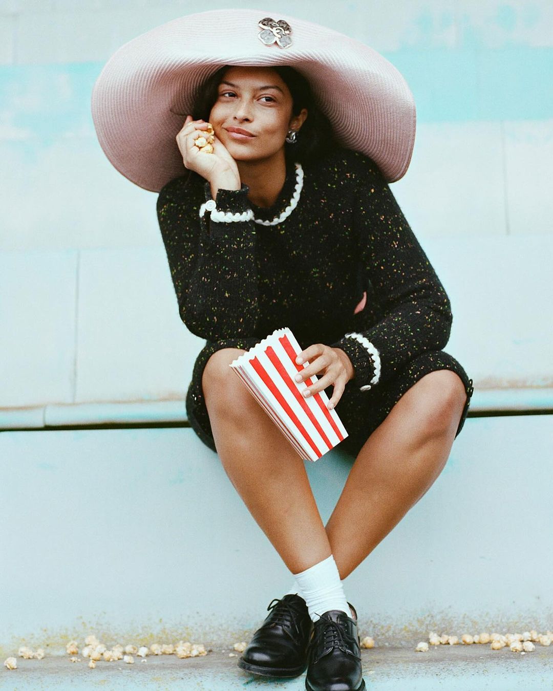 Woman in black dress and oversized hat sits outdoors holding popcorn, creating a whimsical and fashionable portrait with a playful touch.
