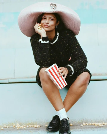Woman in black dress and oversized hat sits outdoors holding popcorn, creating a whimsical and fashionable portrait with a playful touch.