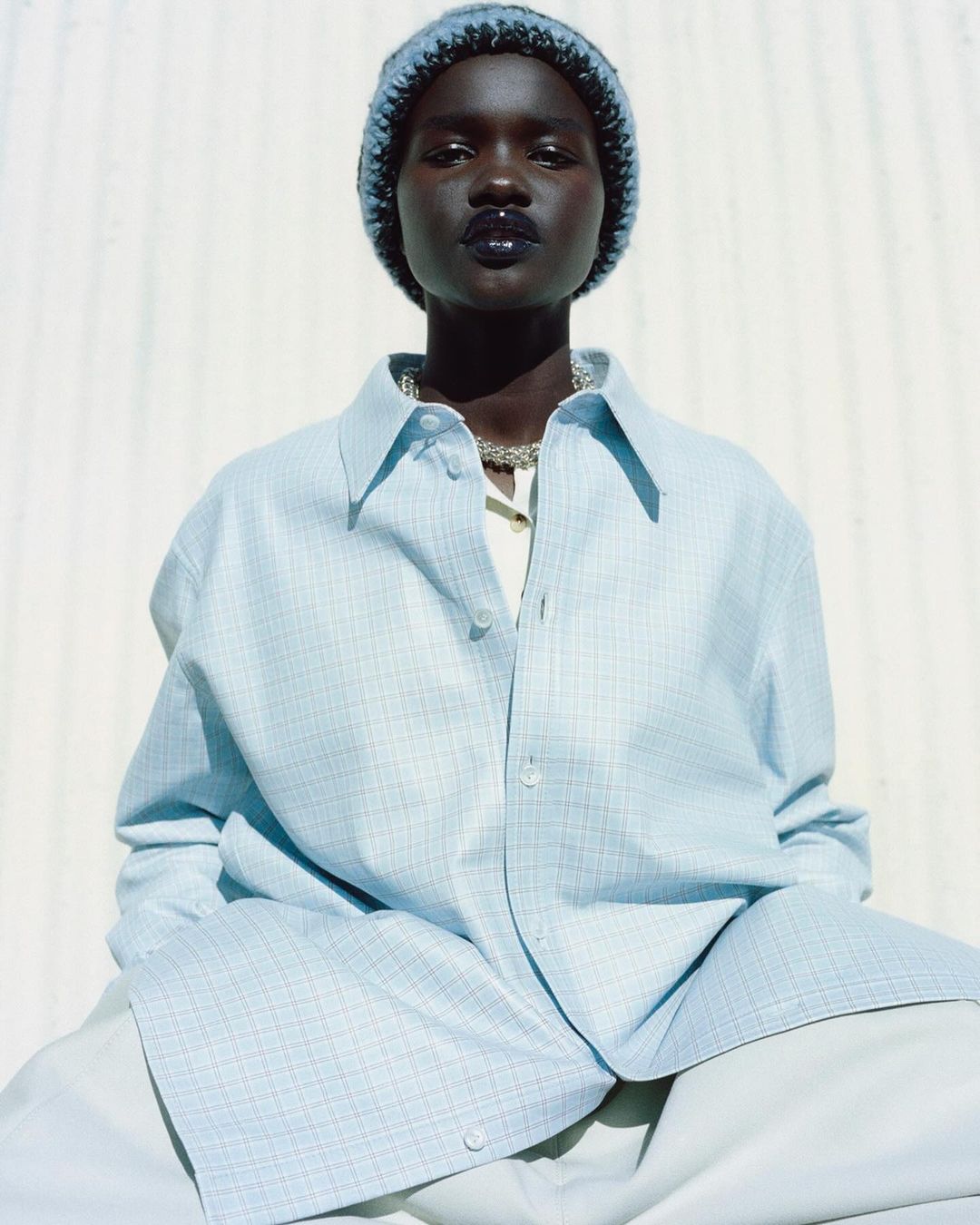 Close-up of a dark-skinned young adult woman with a beanie, silver necklace, and light blue oversized button-down shirt against a white wall.