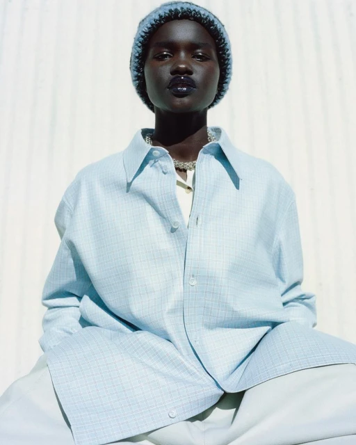 Close-up of a dark-skinned young adult woman with a beanie, silver necklace, and light blue oversized button-down shirt against a white wall.