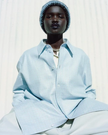 Close-up of a dark-skinned young adult woman with a beanie, silver necklace, and light blue oversized button-down shirt against a white wall.