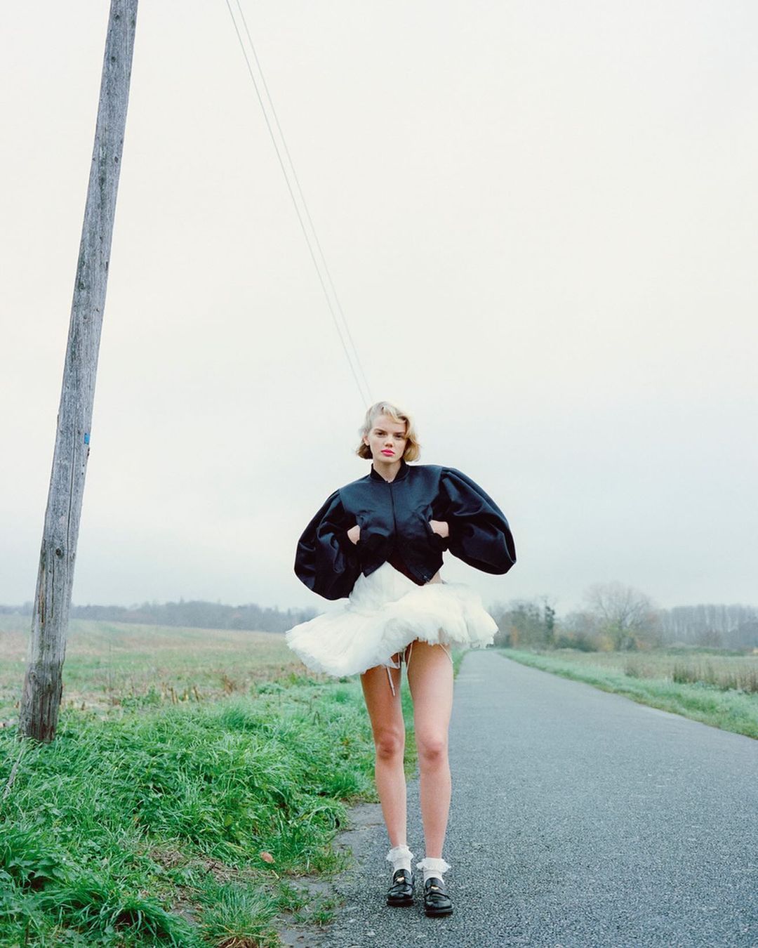 A woman stands in a field wearing a black jacket and white tutu with a rural background, creating an editorial fashion photograph.