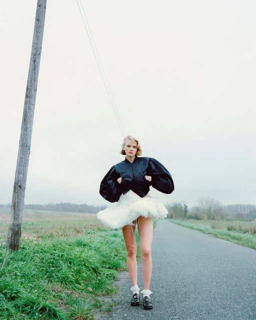 A woman stands in a field wearing a black jacket and white tutu with a rural background, creating an editorial fashion photograph.