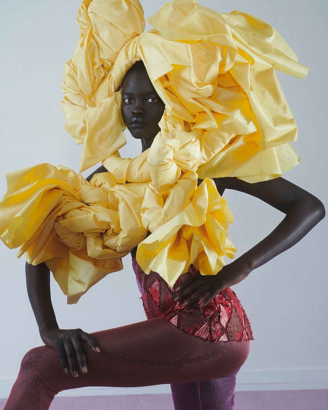 Elegant African woman poses in a striking red top and tights, complemented by a dramatic yellow fabric sculpture around her head and shoulders.