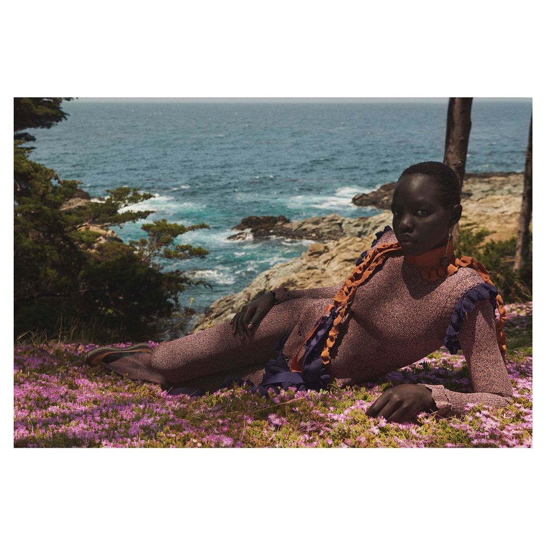 Black model reclining on a flower-covered coastal cliff above the ocean by Julia Noni