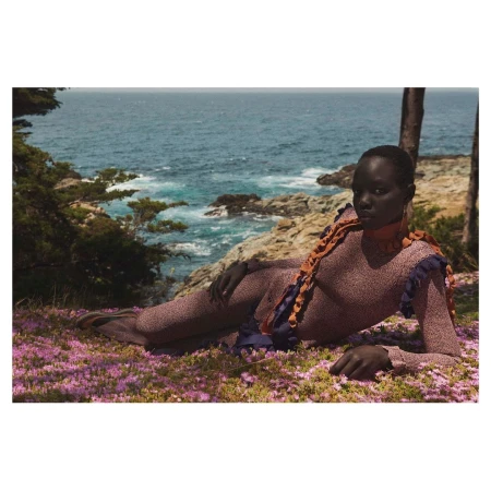 Black model reclining on a flower-covered coastal cliff above the ocean by Julia Noni
