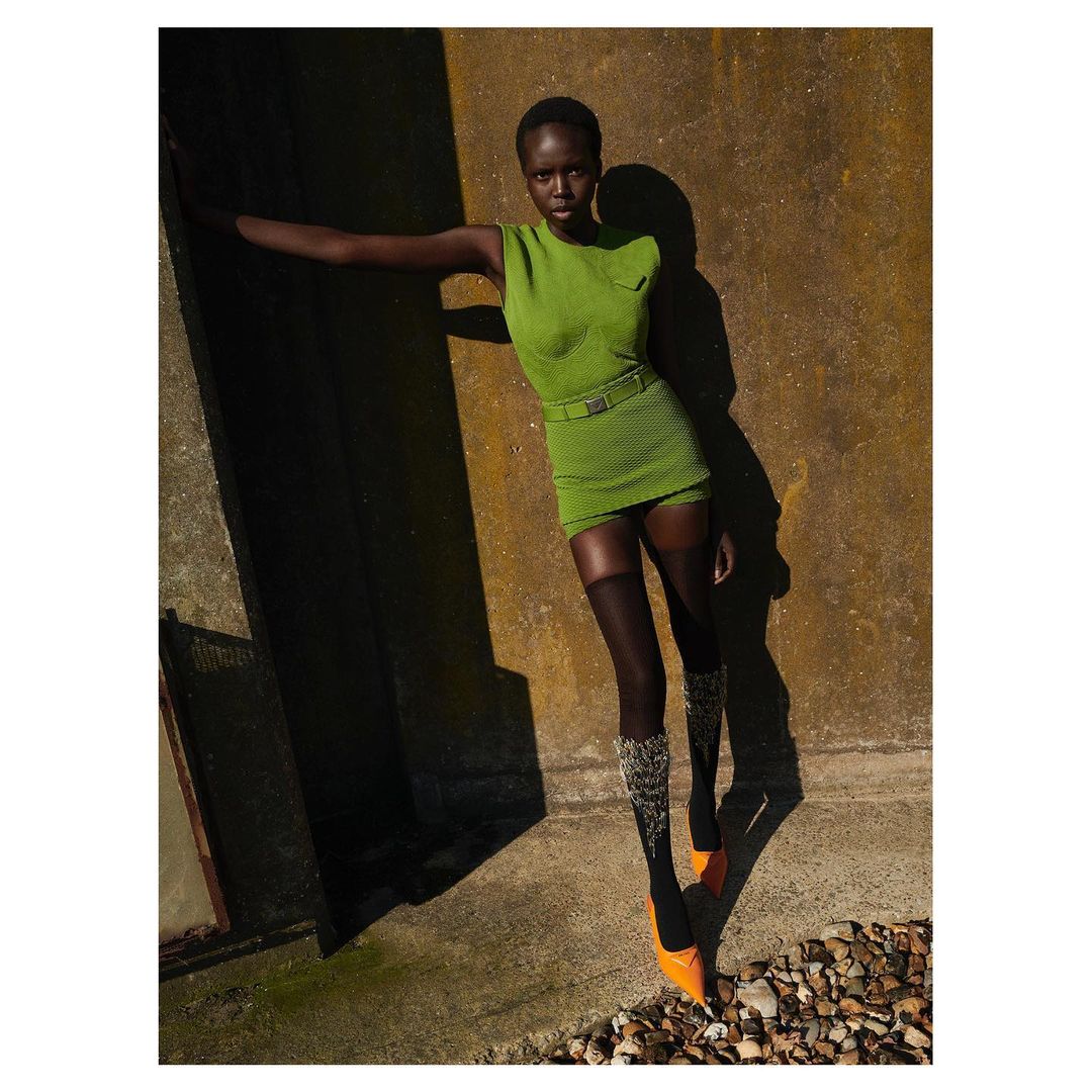 Black woman wearing a green sleeveless dress and orange heels standing against a textured stone wall outdoors.
