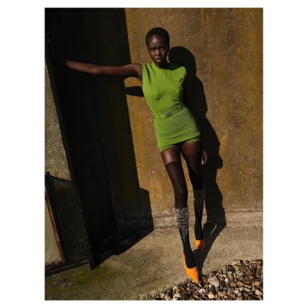 Black woman wearing a green sleeveless dress and orange heels standing against a textured stone wall outdoors.
