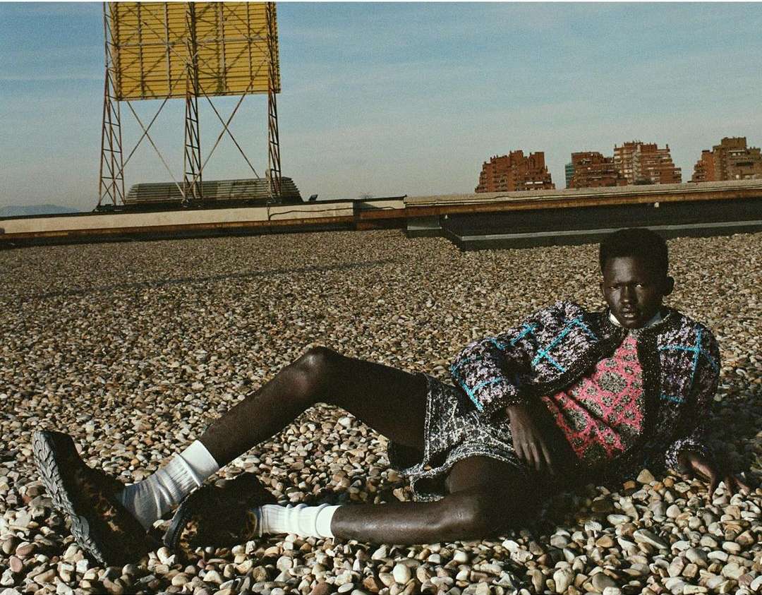 Male model poses on a rooftop, wearing patterned jacket and shorts, set against a blue sky, buildings, and a structural tower.