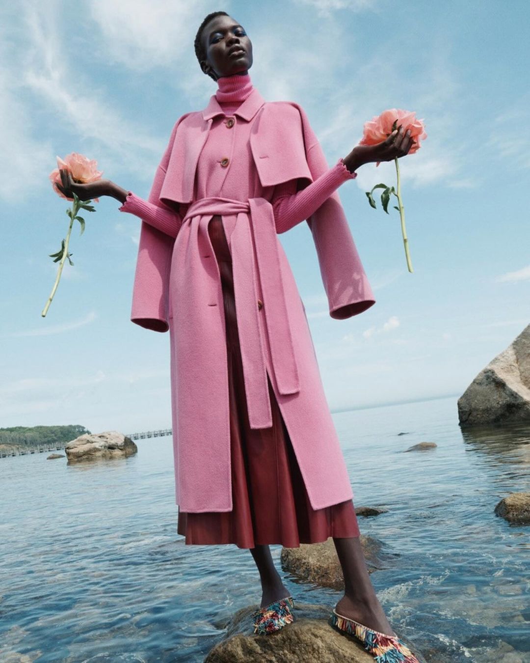 Elegant African woman in pink coat stands on rocks in water holding pink roses against cloudy sky in an outdoor fashion editorial.