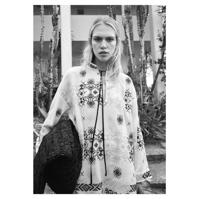 Black and white medium shot of a blonde female adult wearing a patterned white top, holding a dark woven bag, and standing outdoors.