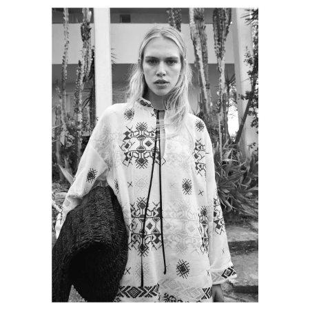 Black and white medium shot of a blonde female adult wearing a patterned white top, holding a dark woven bag, and standing outdoors.