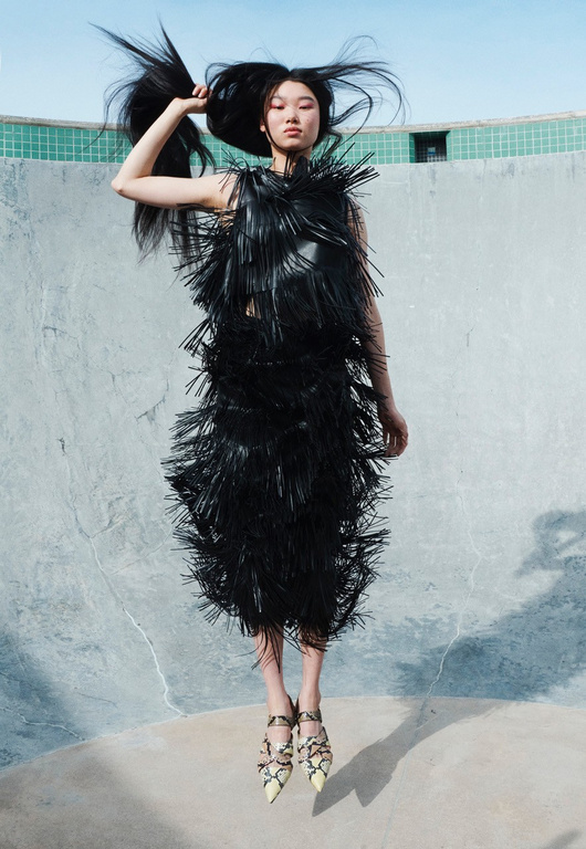 Fashion editorial shot of an Asian model in a black fringe dress, posing dynamically in a concrete structure against a bright sky.