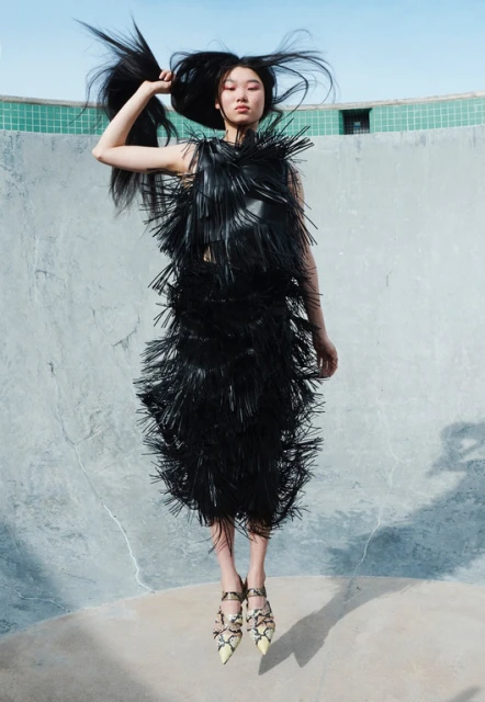 Fashion editorial shot of an Asian model in a black fringe dress, posing dynamically in a concrete structure against a bright sky.