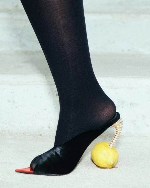 A woman's leg and black shoe with a lemon as a heel rest on a weathered surface in a fashion-forward editorial shot.