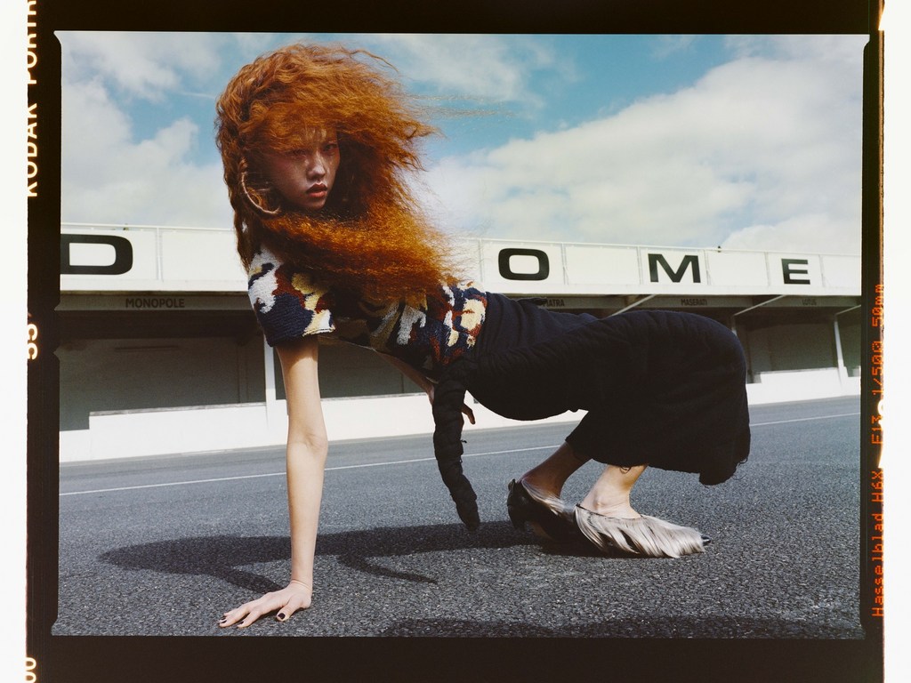 Fashion shot of woman with bright red hair wearing textured top and skirt, posed low on ground with building in the background.