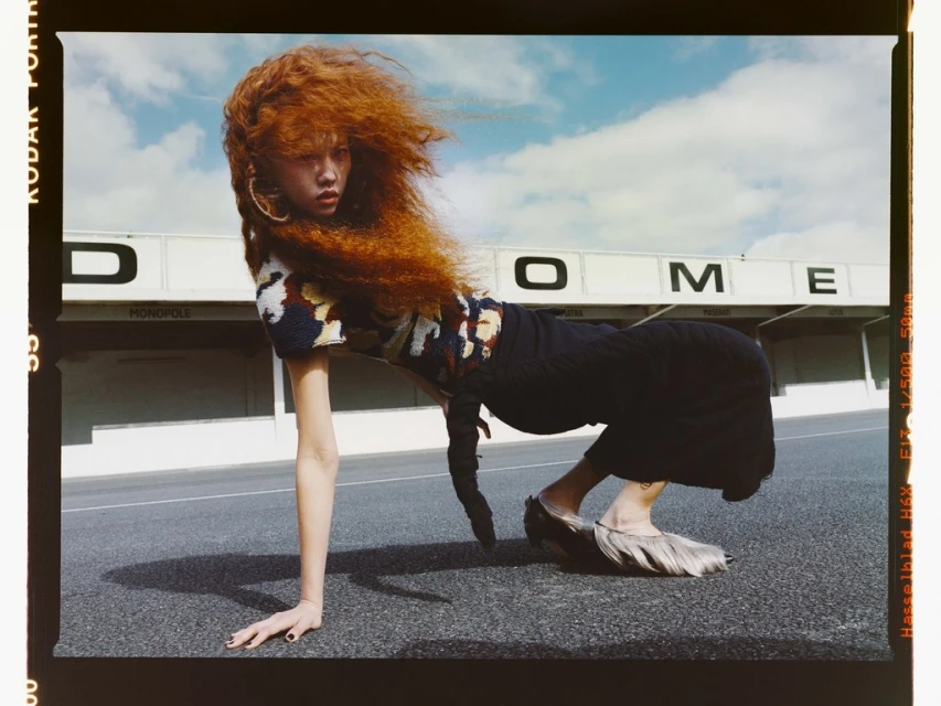 Fashion shot of woman with bright red hair wearing textured top and skirt, posed low on ground with building in the background.