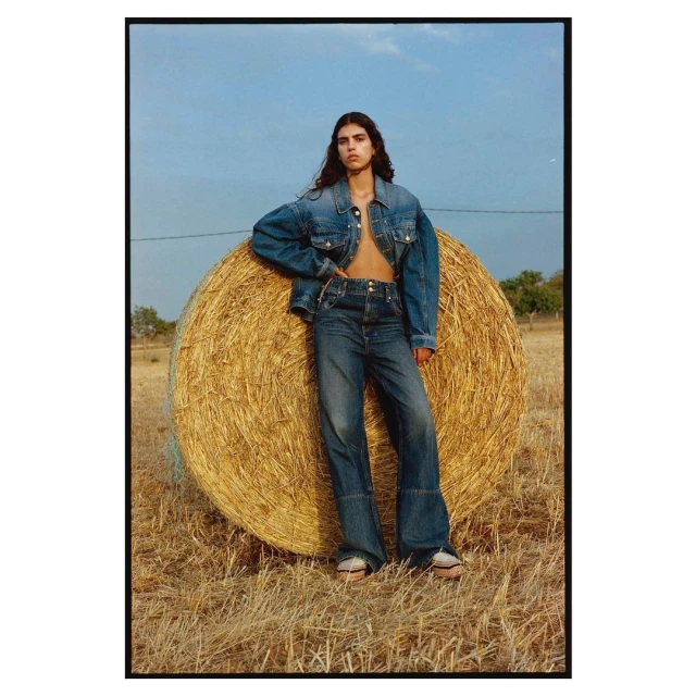 A young woman in denim poses next to a round hay bale in a rural field under a blue sky with a few trees in the background.