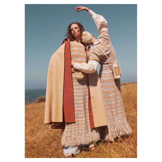 Two models in textured dresses and coat pose in a field with blue sky and ocean backdrop, fashion-forward editorial style.