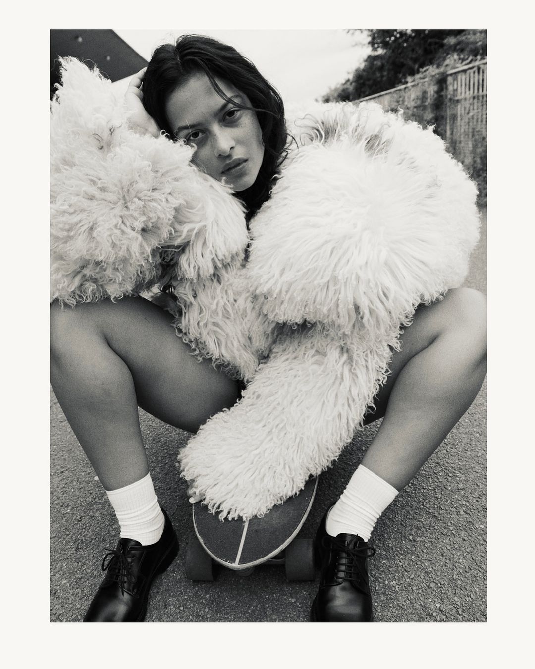 Monochrome portrait of a woman wearing a fluffy textured coat crouched on a skateboard with white socks and dark shoes