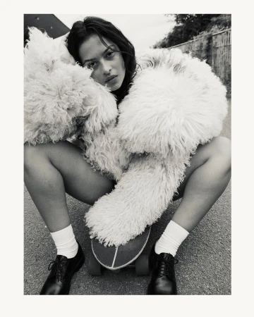Monochrome portrait of a woman wearing a fluffy textured coat crouched on a skateboard with white socks and dark shoes