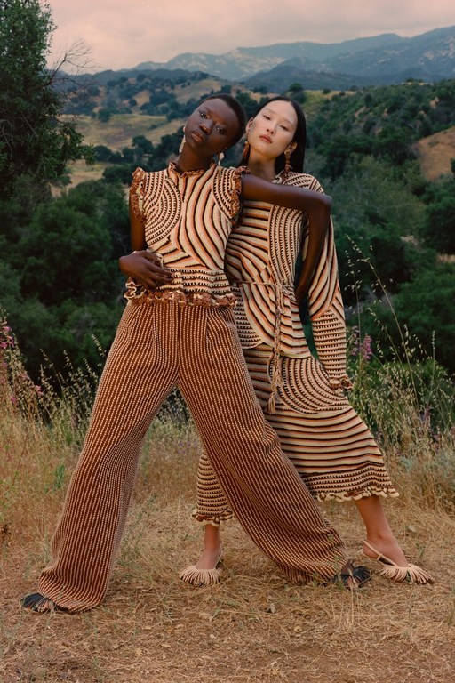 Two women model striped knitwear in an outdoor setting with rolling hills. A darker-skinned woman stands beside a lighter-skinned woman.