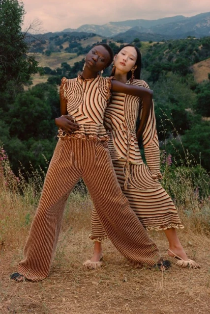 Two women model striped knitwear in an outdoor setting with rolling hills. A darker-skinned woman stands beside a lighter-skinned woman.